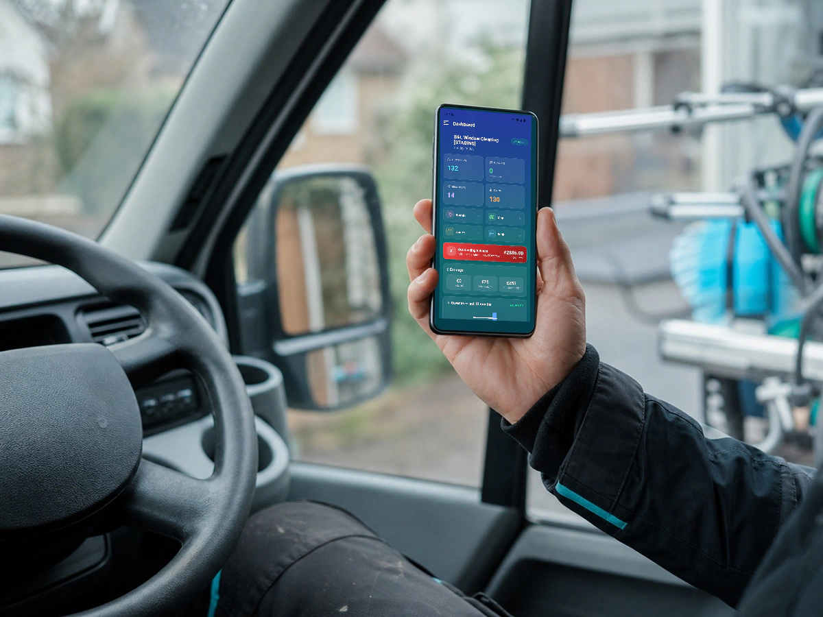Window cleaner holding a phone displaying the RoundRun dashboard inside a work van